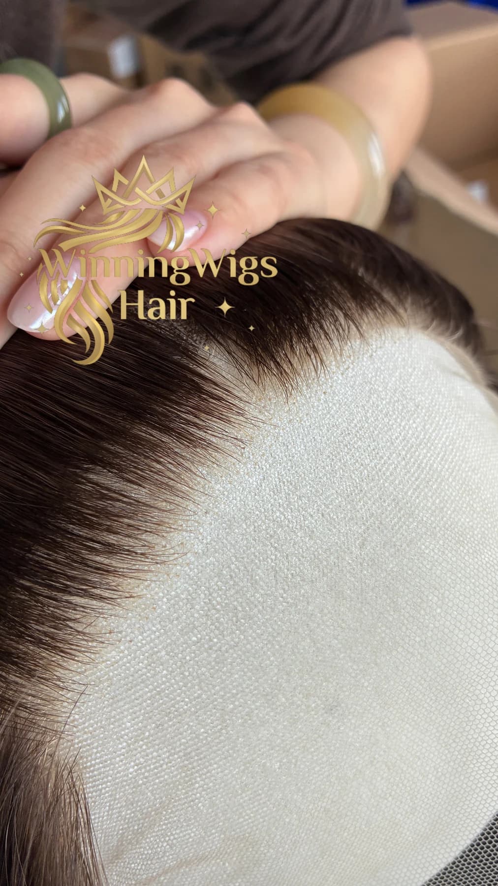 Close-up of a brown wig hairline and lace on a white textured backing while a hand holds the hair away from the front