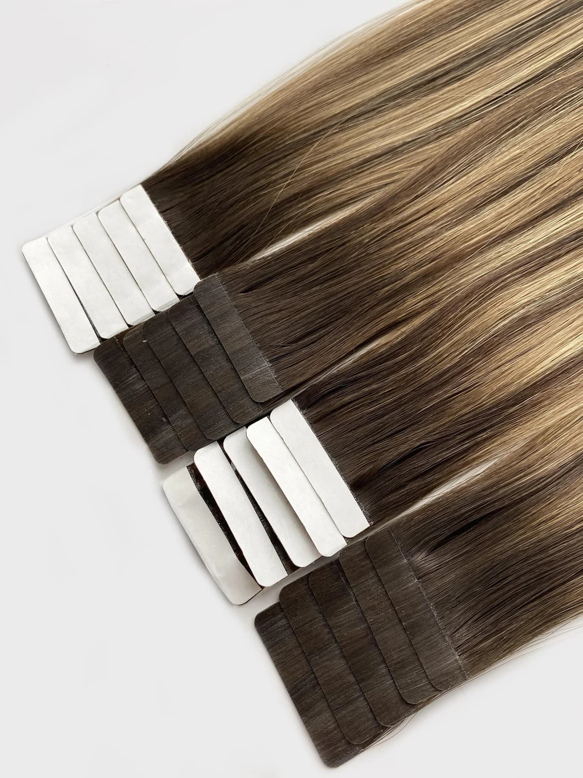 Close-up of rooted brown and blonde tape-in hair extensions with white protective liners and dark attachment bases