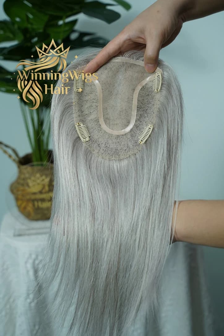 Underside of a gray hair topper held open to show its base, beige edging and four attachment clips